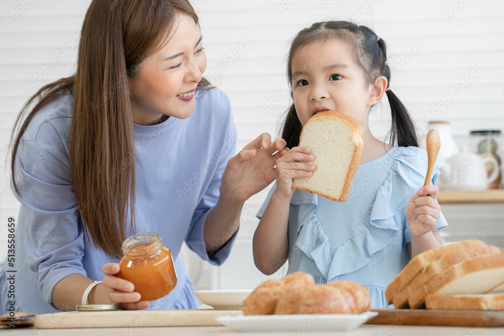 Happy Asian family, cute little daughter eating bread with jam and young mother looking at her baby with love after spreading jam on sliced bread together, preparing breakfast in kitchen at home Happy Asian family, cute little daughter eating bread with jam and young mother looking at her baby with love after spreading jam on sliced bread together, preparing breakfast in kitchen at home