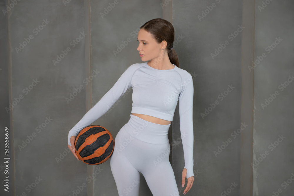 Young fit athletic woman in white sportswear holding a basketball ball, looking ahead and standing on a grey stone wall background.

Sporty woman taking break after workout or basketball game.