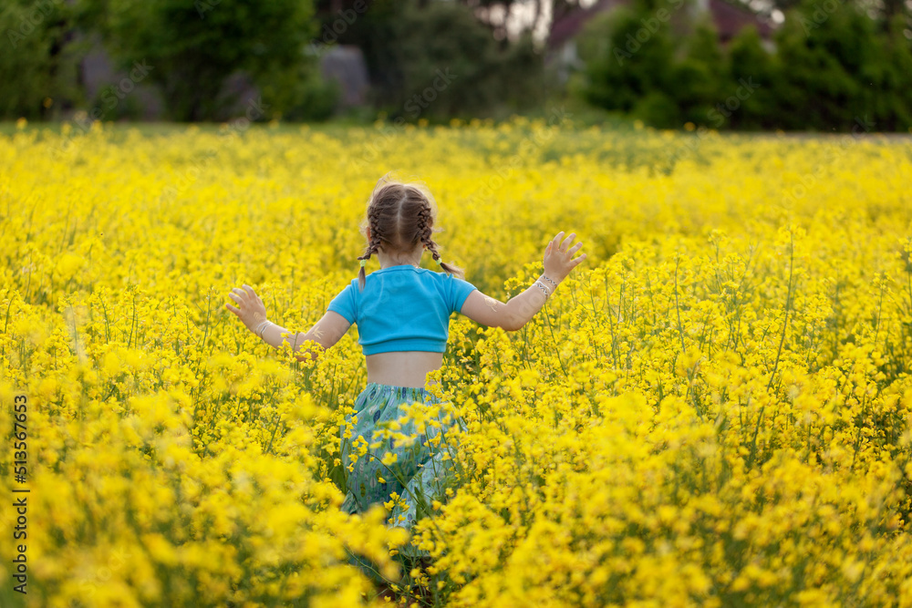 Cute little girl running through yellow rape flowewrs field. Girl in blue dress hands raised to side enjoys nature having fun jogging through meadow at sunny day. Back view. Cute little girl running through yellow rape flowewrs field. Girl in blue dress hands raised to side enjoys nature having fun jogging through meadow at sunny day. Back view.