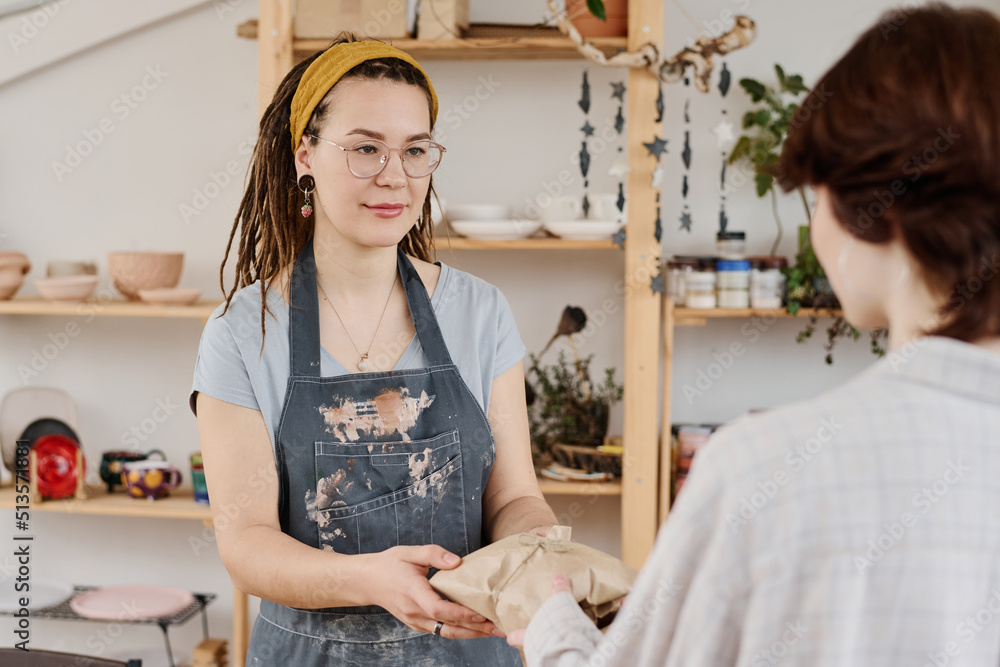 Young female seller in workwear looking at buyer while passing her wrapped handmade earthenware product in small shop Young female seller in workwear looking at buyer while passing her wrapped handmade earthenware product in small shop
