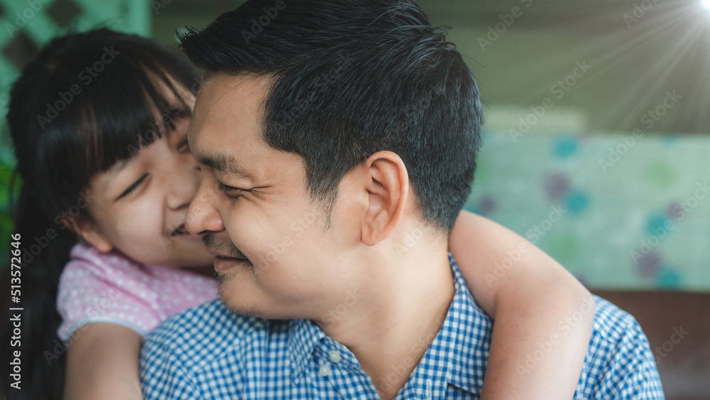 Father and daughter are hugging with love, looking at camera and smiling while sitting on sofa at home.Concept of Father's day
