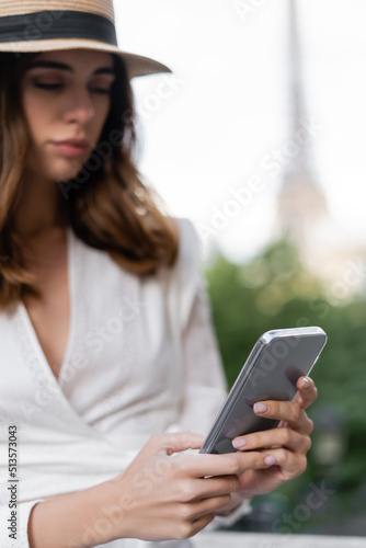 Blurred young woman in sun hat using smartphone outdoors in Paris.