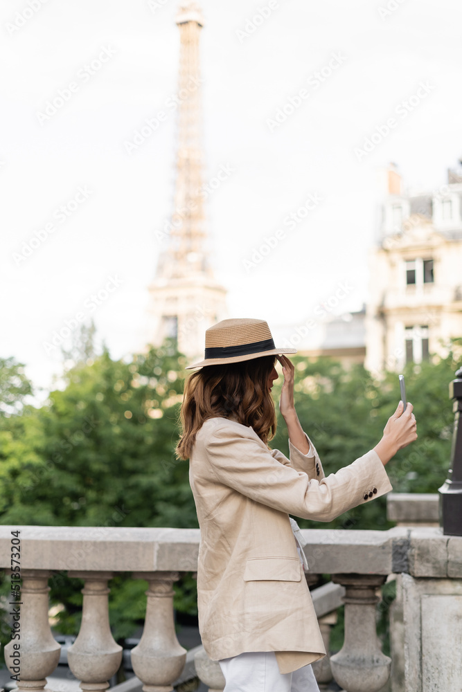 Side view of stylish woman in sun hat taking selfie on smartphone in Paris.