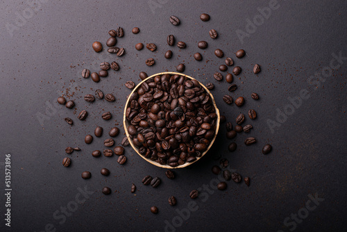 A bowl is full of coffee beans on a black surface with scattered coffee beans. Aerial view plan.