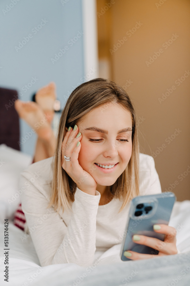 Young white blond woman smiling and using cellphone in bed at home Young white blond woman smiling and using cellphone in bed at home