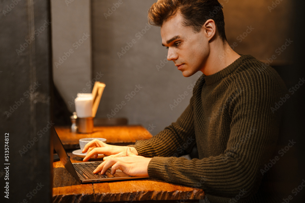 Young man wearing sweater using laptop and drinking coffee in cafe Young man wearing sweater using laptop and drinking coffee in cafe