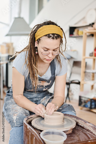 Creative woman in casualwear sitting by rotating pottery wheel in workshop and creating new clay items for selling in earthenware shop