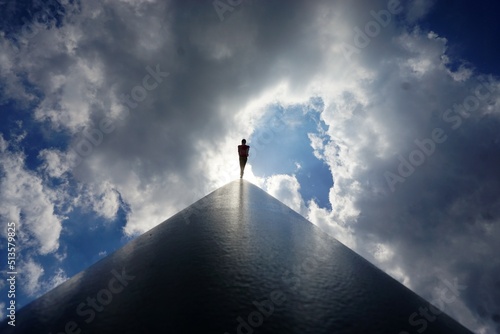 Architektonisches Bauwerk aus Stahlbeton mit emporragendem Pfahl und Person vor Wolkengebilde am Himmel 