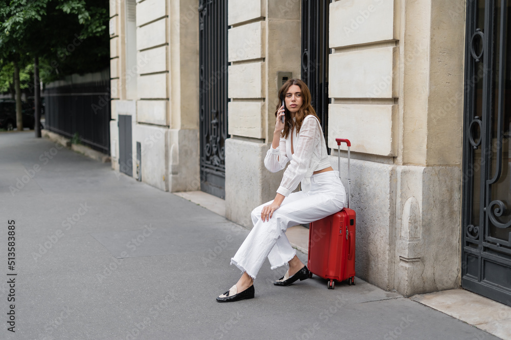 Young woman talking on smartphone while sitting on suitcase on urban street in Paris. Young woman talking on smartphone while sitting on suitcase on urban street in Paris.