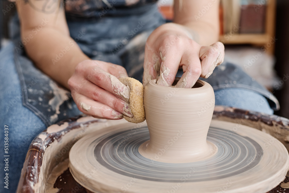 Hand of female potter holding sponge close to rotating clay item on pottery wheel while creating new earthenware in workshop Hand of female potter holding sponge close to rotating clay item on pottery wheel while creating new earthenware in workshop