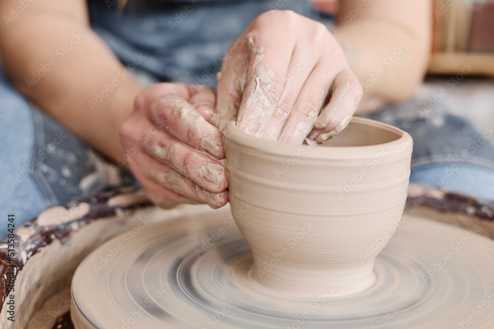Hands of female owner of small earthenware shop creating new pot, bowl or other clay item while sitting in front of rotating pottery wheel Hands of female owner of small earthenware shop creating new pot, bowl or other clay item while sitting in front of rotating pottery wheel