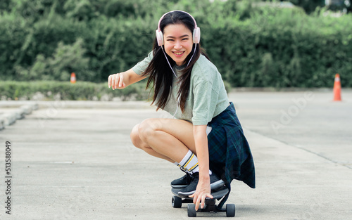 Beautiful sportive hipster happy Asian woman wearing casual shirt and shorts with headphone to listen music while playing skateboard as hobby in free time day on holiday, smiling at outdoor park.