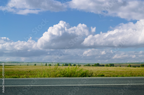 Majestic clouds over an asphalt road, bushes and a field with trees