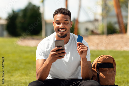 Fototapeta Naklejka Na Ścianę i Meble -  Promo code for purchase or online shopping. Happy black man using smartphone and credit card, sitting in urban city area