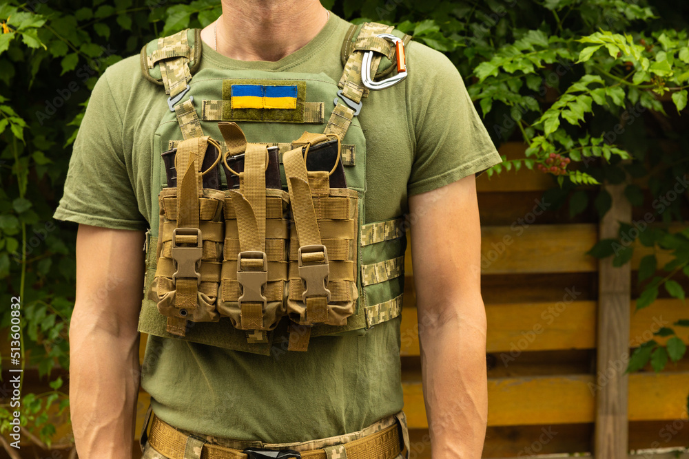 A soldier in a camouflage tactical uniform and a military body armor on a  background of wood and greenery. Body armor with combat butts. Ukrainian  army. Military concept. No war.medical tourniquet. Stock