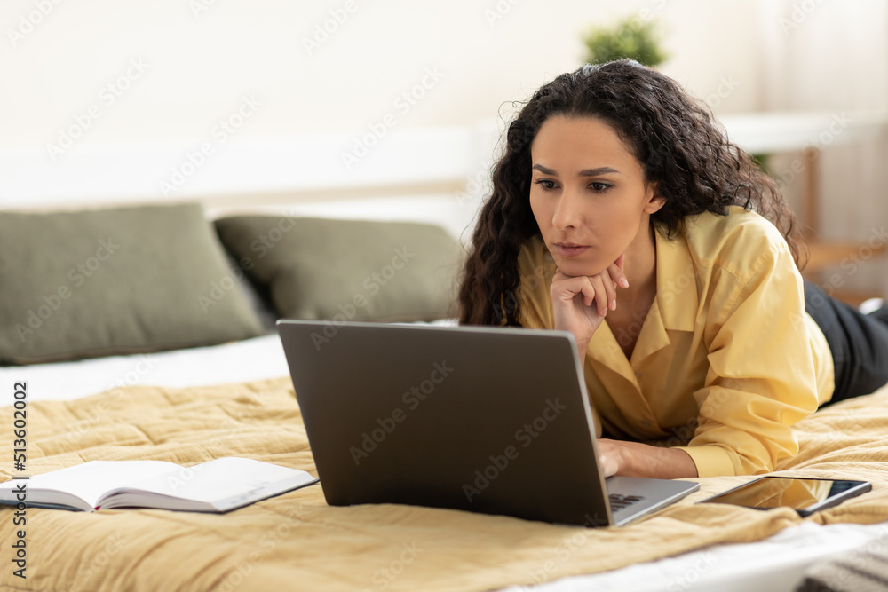 Serious young woman using laptop, working or studying online, having business meeting or webinar, lying on bed at home Serious young woman using laptop, working or studying online, having business meeting or webinar, lying on bed at home