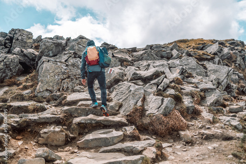 Young traveler man in sportswear with a backpack climbs the rocky road up to the top of the mountain in summer. Black view