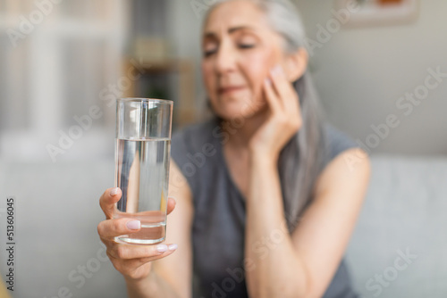 Unhappy sad european old woman with gray hair hold glass of water, suffers from pain