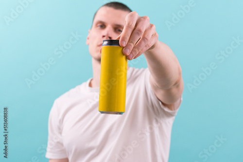 A man in a white t-shirt holds a yellow empty aluminum can on a blue background. Metallic. Mock. Alcohol. Design. Blank. White. Water. Cold. Liquid. Product. Mockup. Tinned. Refresh. Label. Empty