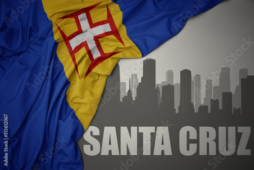 abstract silhouette of the city with text Santa Cruz near waving national flag of madeira on a gray background.