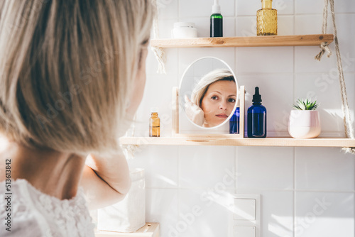 Beautiful middle-aged woman looking in the mirror in white eco friendly bathroom. Wooden shleves and reusable cosmetics bottles. Wellnes concept