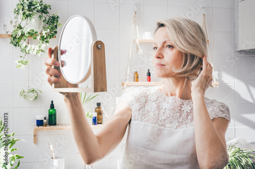 Middle-aged woman looking in the mirror in light bathroom with biophilic minimalistic design. Aging skin care. Wellnes