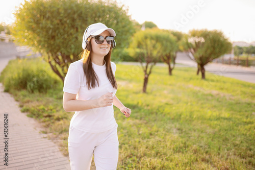 A female runner in white clothes runs at sunset in the summer, listening to music in headphones.