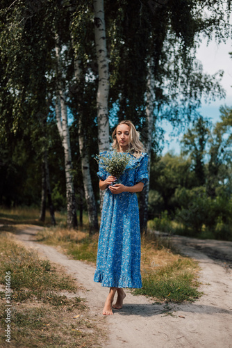 A young beautiful woman in a blue dress holds wild flowers in her hands
