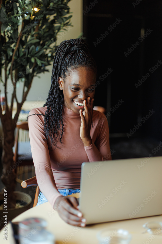Portrait of young smiling African-American woman with long dark braids wearing pink roll-neck, sitting at table in cafe near laptop, looking at screen, leaning head on hand. Technology, videocall. Portrait of young smiling African-American woman with long dark braids wearing pink roll-neck, sitting at table in cafe near laptop, looking at screen, leaning head on hand. Technology, videocall.