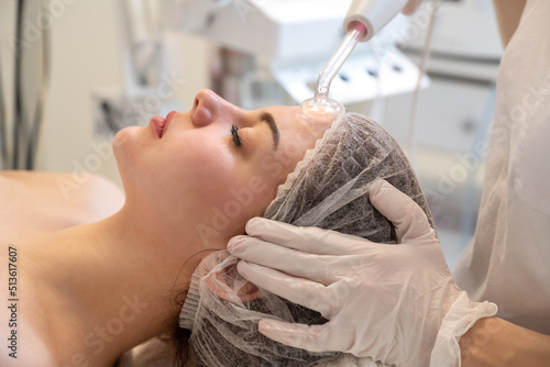 Beautician applying electric Darsonval facial massage treatment with special tool over female patients forehead in beauty salon photo