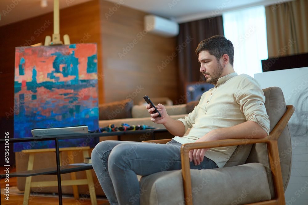 Modern young guy digital artist holding phone checking NFT sales sitting in creative studio, workshop. Remote work