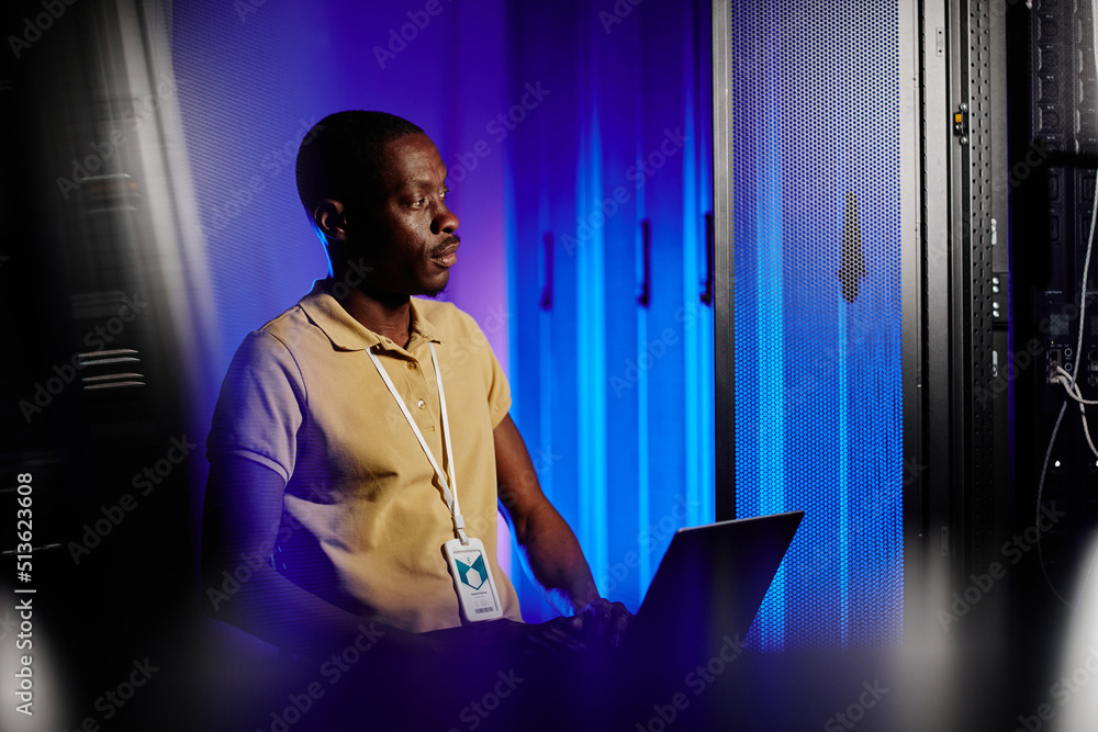 © Seventyfour - Side view portrait of adult black man using laptop in dark server room lit by neon light, copy space © Seventyfour - Side view portrait of adult black man using laptop in dark server room lit by neon light, copy space