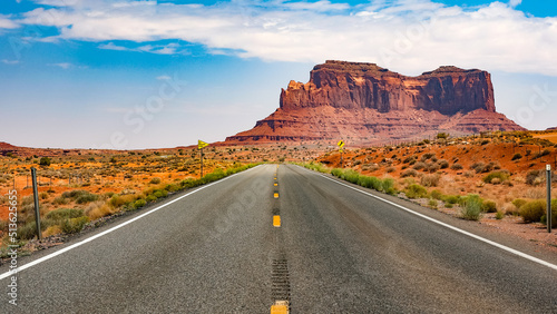 A road stretches to the horizon. A mesa rises in the distance. Monument Valley, Utah.