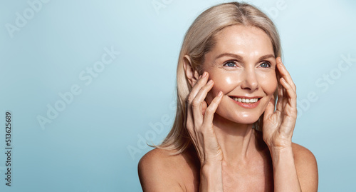 Smiling middle aged woman, 50 years model, applies anti-aging skin cream, rubbing in moisturizer on face, standing over blue background photo