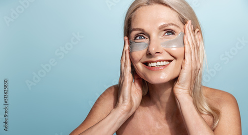 Smiling elderly woman with no blemishes, perfect skin, usng under eye patches from puffiness, wrinkle reduction and dark circles, blue background