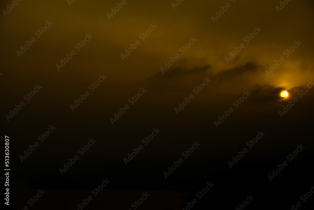 Moody sky with dark clouds and a deep orange sun in a stormy setting at sunset. Light illuminates clouds creating an eerie and ominous atmosphere.