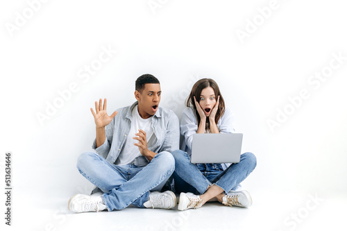 Amazed stunned young multiracial couple, in casual wear, sitting on floor over isolated white background with laptop, looking at a screen in surprise, saw unexpected news, big discount in online store