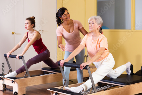 Friendly Hispanic woman professional pilates instructor assisting elderly female to do exercises on reformer. Active lifestyle concept