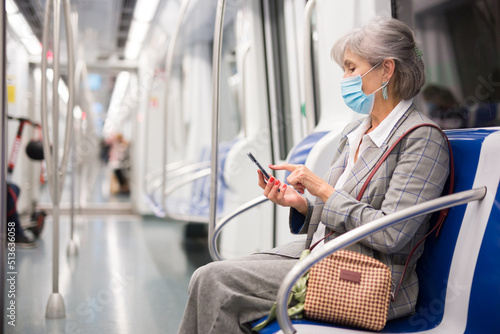 Caucasian mature woman in face mask sitting inside subway train and using her smartphone.