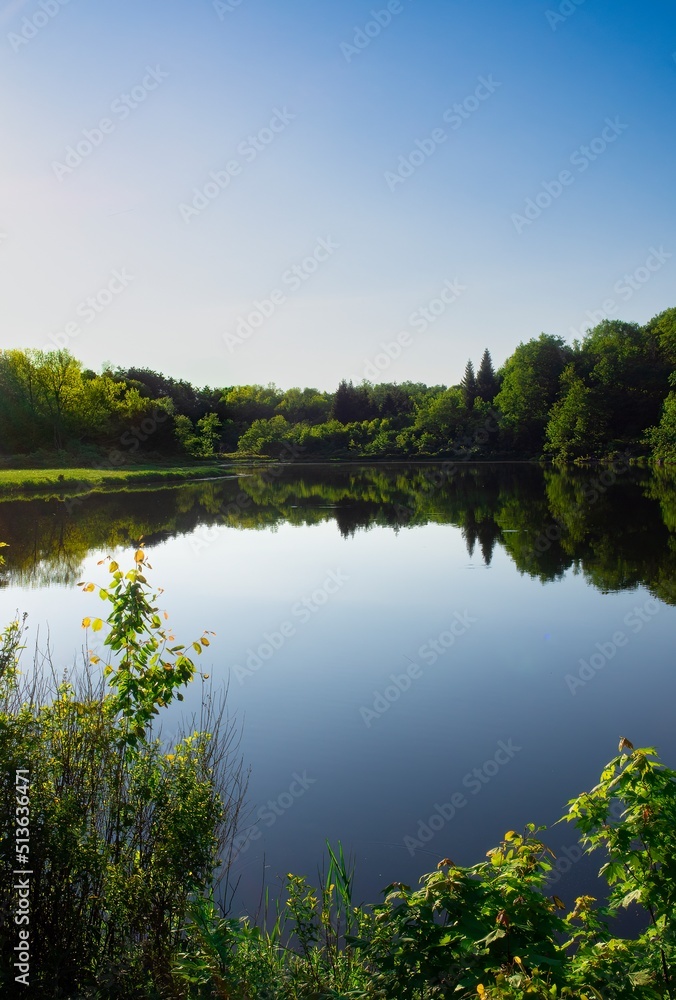 Small lake or pond, mirror-like, calm surface, in late afternoon. Quiet, subdued mood. Reflections of trees surrounding the lake in the water.