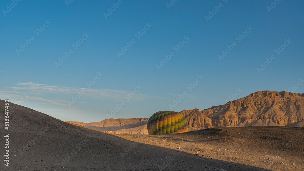 A bright balloon rises from the gorge above the desert plateau.  Shadows on the sand. A picturesque mountain range against a clear blue sky. Copy space. Egypt. Luxor