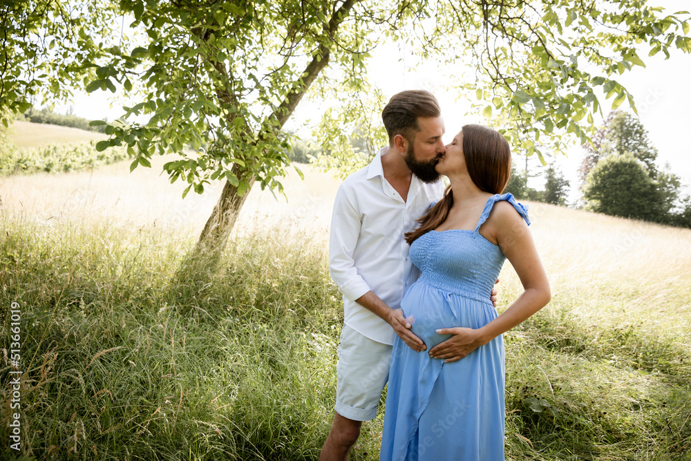 Naklejka premium young couple in love standing in high flower meadow in summer and cuddling and the woman is pregnant and has a blue dress on and the man has a full beard