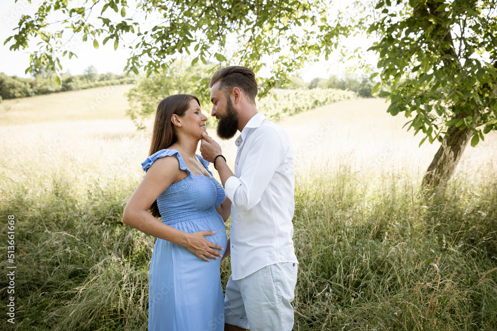 Fototapeta premium young couple in love standing in high flower meadow in summer and cuddling and the woman is pregnant and has a blue dress on and the man has a full beard