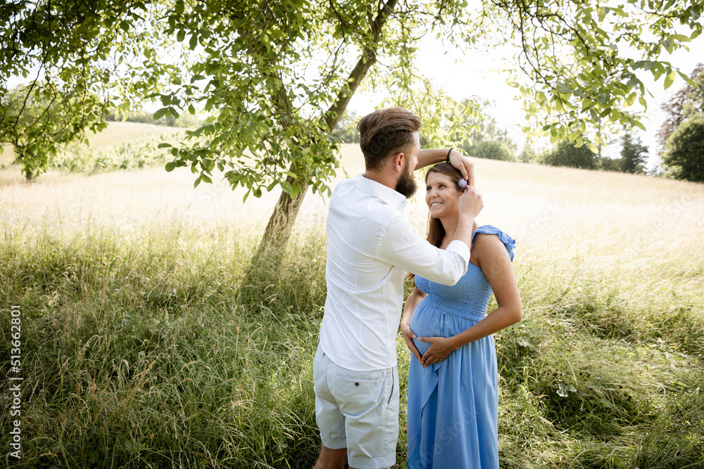 young couple in love standing in high flower meadow in summer and cuddling and the woman is pregnant and has a blue dress on and the man has a full beard young couple in love standing in high flower meadow in summer and cuddling and the woman is pregnant and has a blue dress on and the man has a full beard