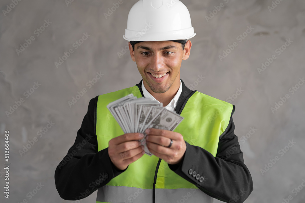 Young arabic builder man engineer wearing safety helmet holding dollars as payment over background with raised hand, payment construction Young arabic builder man engineer wearing safety helmet holding dollars as payment over background with raised hand, payment construction