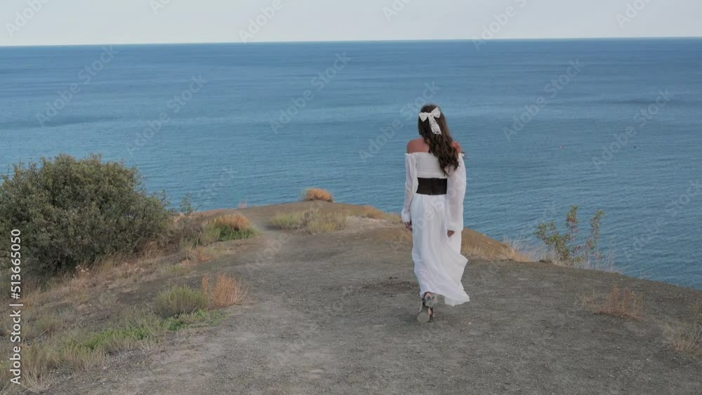 A young beautiful girl with dark hair, dressed in a white dress, walks down the slope at the top of the mountain, the wind blows her hair and dress, and smiles - harmony, adventure, freedom, 4k video A young beautiful girl with dark hair, dressed in a white dress, walks down the slope at the top of the mountain, the wind blows her hair and dress, and smiles - harmony, adventure, freedom, 4k video
