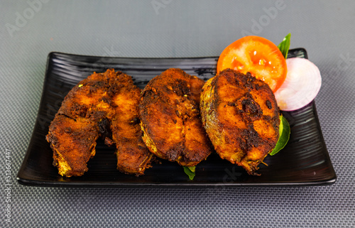 Tasty fish fry arranged beautifully and garnished with onion, tomato slices and green leaves on a black ceramic plate with a grey textured background.