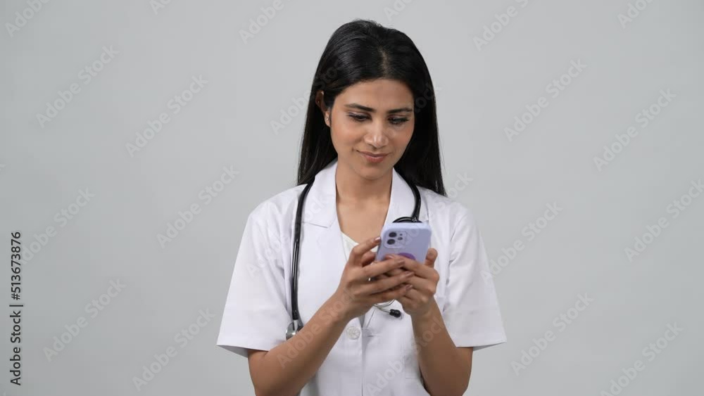 Happy young Indian woman doctor wearing white medical coat and stethoscope,chatting on smartphone. Smiling Asian female physician posing in studio. Happy young Indian woman doctor wearing white medical coat and stethoscope,chatting on smartphone. Smiling Asian female physician posing in studio.
