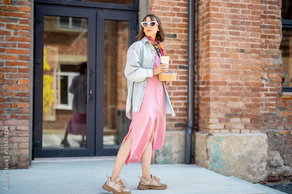 Young stylish woman walks with coffee cup near modern office building outdoors. Portrait of female hipster during a coffee break