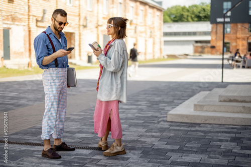 Young stylish couple have some conversation while standing together with phones during a coffee break. Young hipsters hang out together near office outdoors